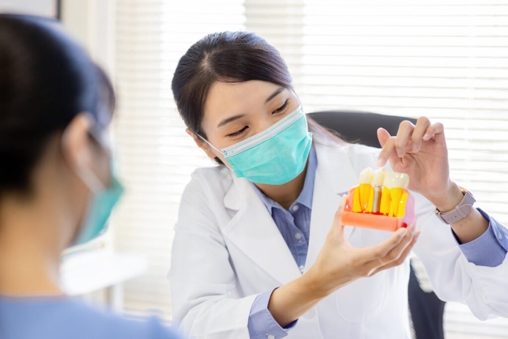 close up of asian female dentist introduces patient for dental implant by denture model while they wear protective face mask to prevent COVID19